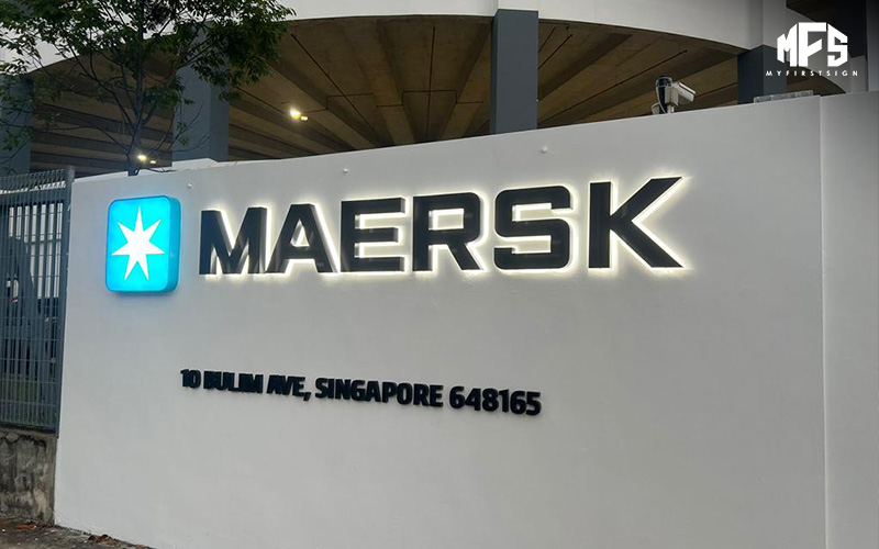 A backlit MAERSK sign with a blue star icon on a white wall at an address in Singapore.